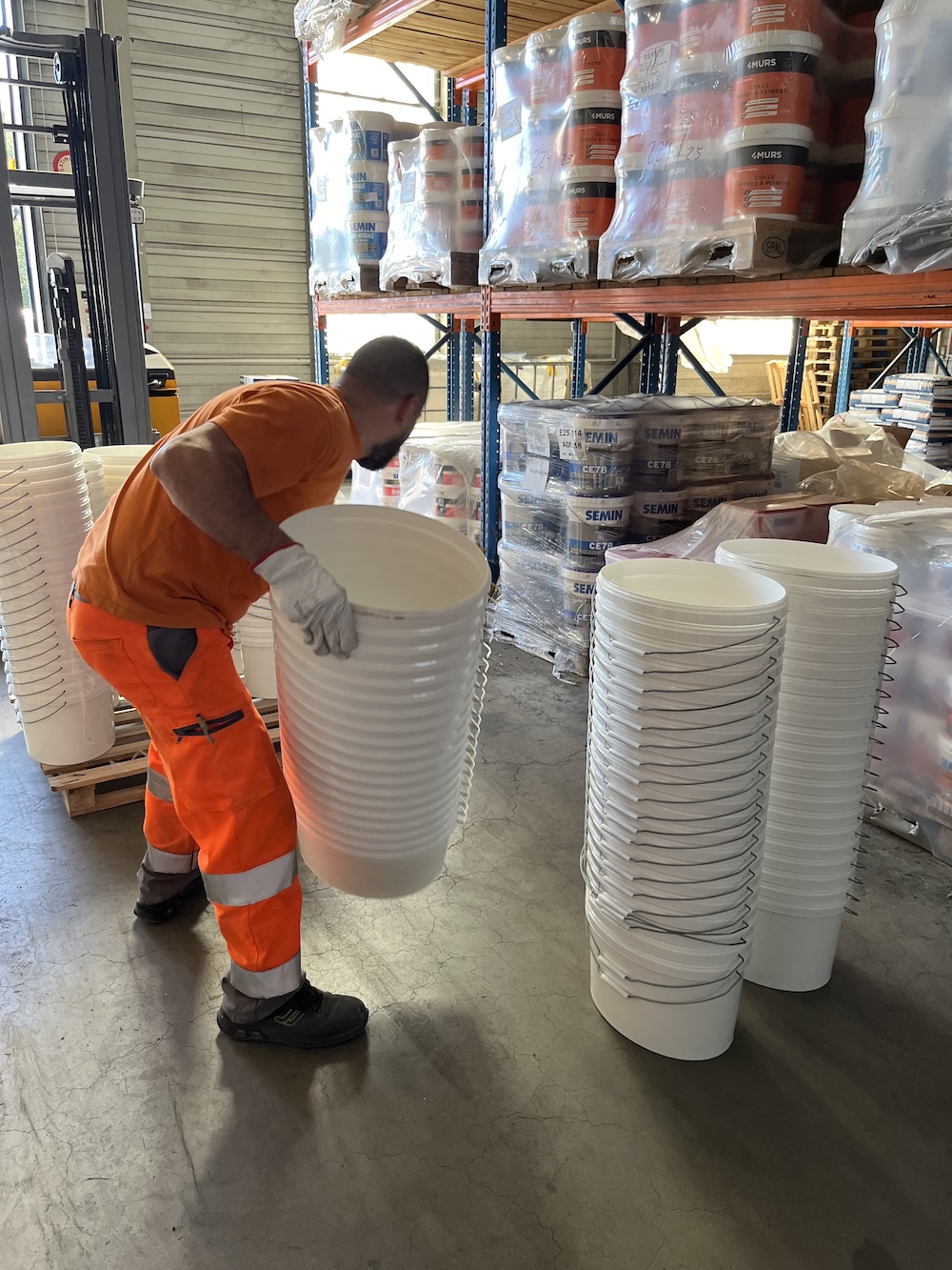 opérateur logistique manipulant des seaux de peinture dans un entrepôt industriel